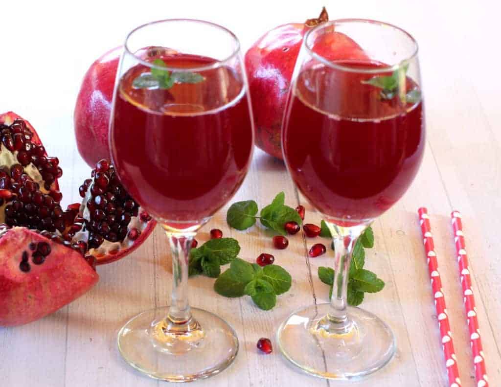 pomegranate juice in 2 wine glasses, with 2 starws on the side and a few pomegranates and mint leaves next to the glasses