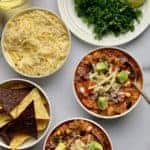 2 white bowls of buffalo chicken chili garnished with cheese and avocados, bowl of tortilla chips, cheese and a plate with chopped cilantro and scallions