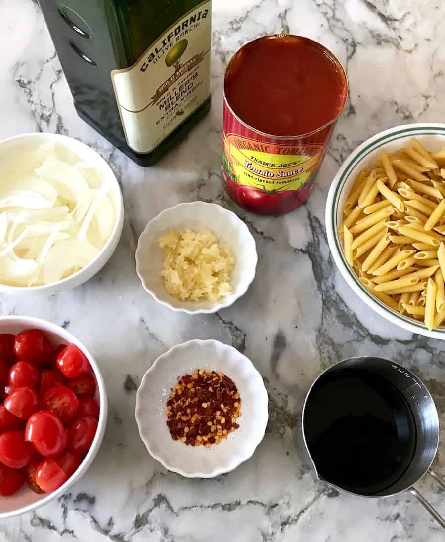 ingredients - olive oil, sliced onions, minced garlic, tomato sauce, sliced grape tomatoes, red chili flakes, dry pasta in a bowl and balsamic vinegar in a measuring cup.
