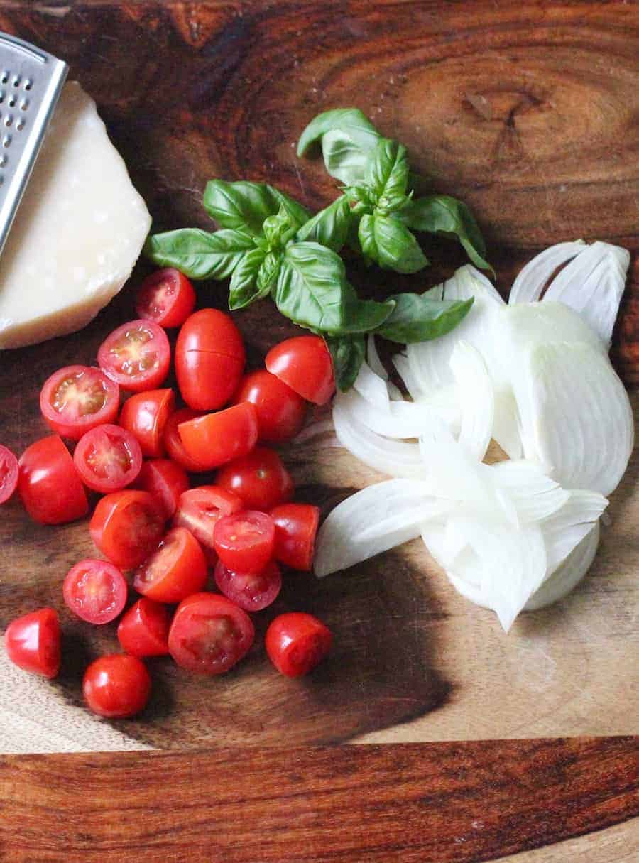 Wooden cutting board with sliced onions, halved grape tomatoes, basil leaves, parmesan cheese and ga rater.