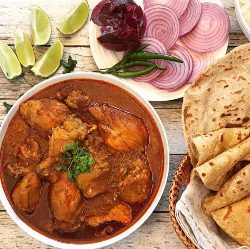 Chicken Curry in a bowl with rotis, lime wedges and sliced onions 