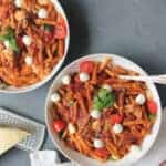 2 white bowls with pasta caprese garnished with mozzarella balls and basil. Parmesan cheese block and cheese grater