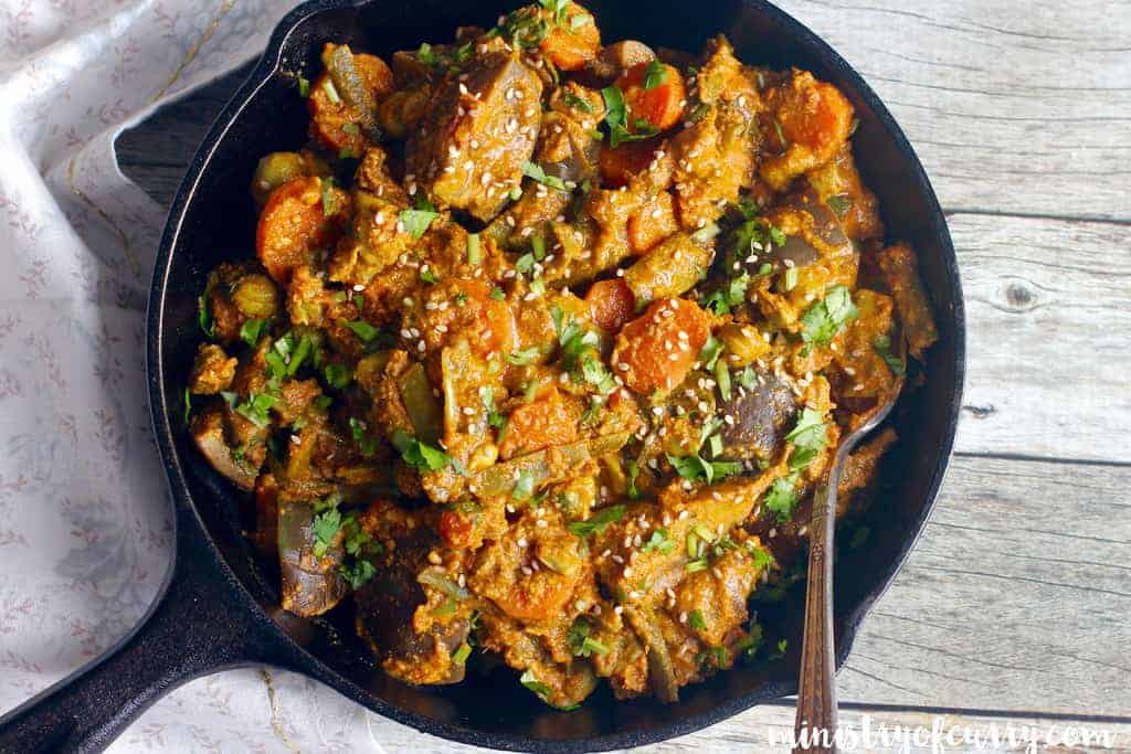 mixed vegetable curry with sesame sauce in a black skillet garnished with sesame seeds and cilantro with a spoon in it. 
