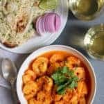 Shrimp curry in a white serving bowl, a white plate with cut up naan, sliced red onions and lemon wedges. Sparking drink in 2 cups on the side.