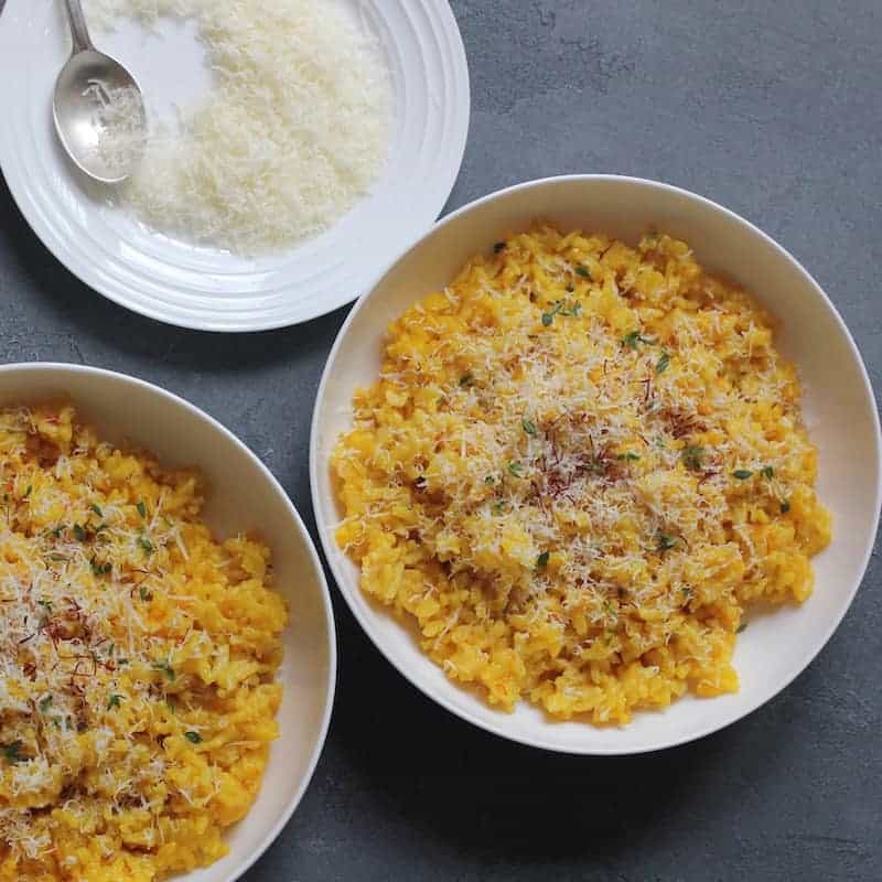 Saffron Risotto, garnished with parmesan cheese and saffron served in 2 white bowls. A plate of grated parmesan cheese on the side with a spoon. 