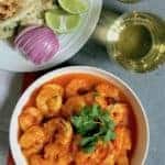 Shrimp curry in a white serving bowl, a white plate with cut up naan, sliced red onions and lemon wedges. Sparking drink in 2 cups on the side.