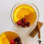 2 cups with spiced apple cider, a wedge of orange and 4 cranberries floating on top. Orange slices, cinnamon sticks and cloves next to the cups on the table.