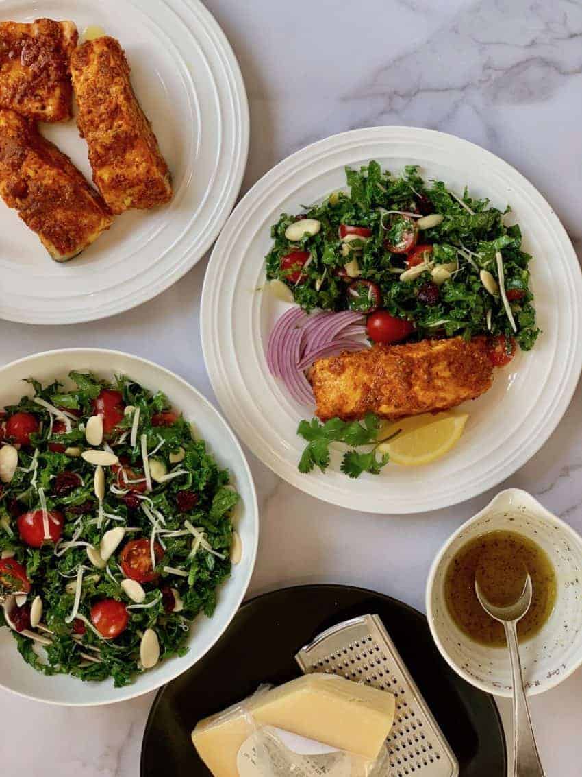 Tandoori Salmon served with kale salad in a white plate. Bowl of salad and a platter with tandoori salmon on the side 