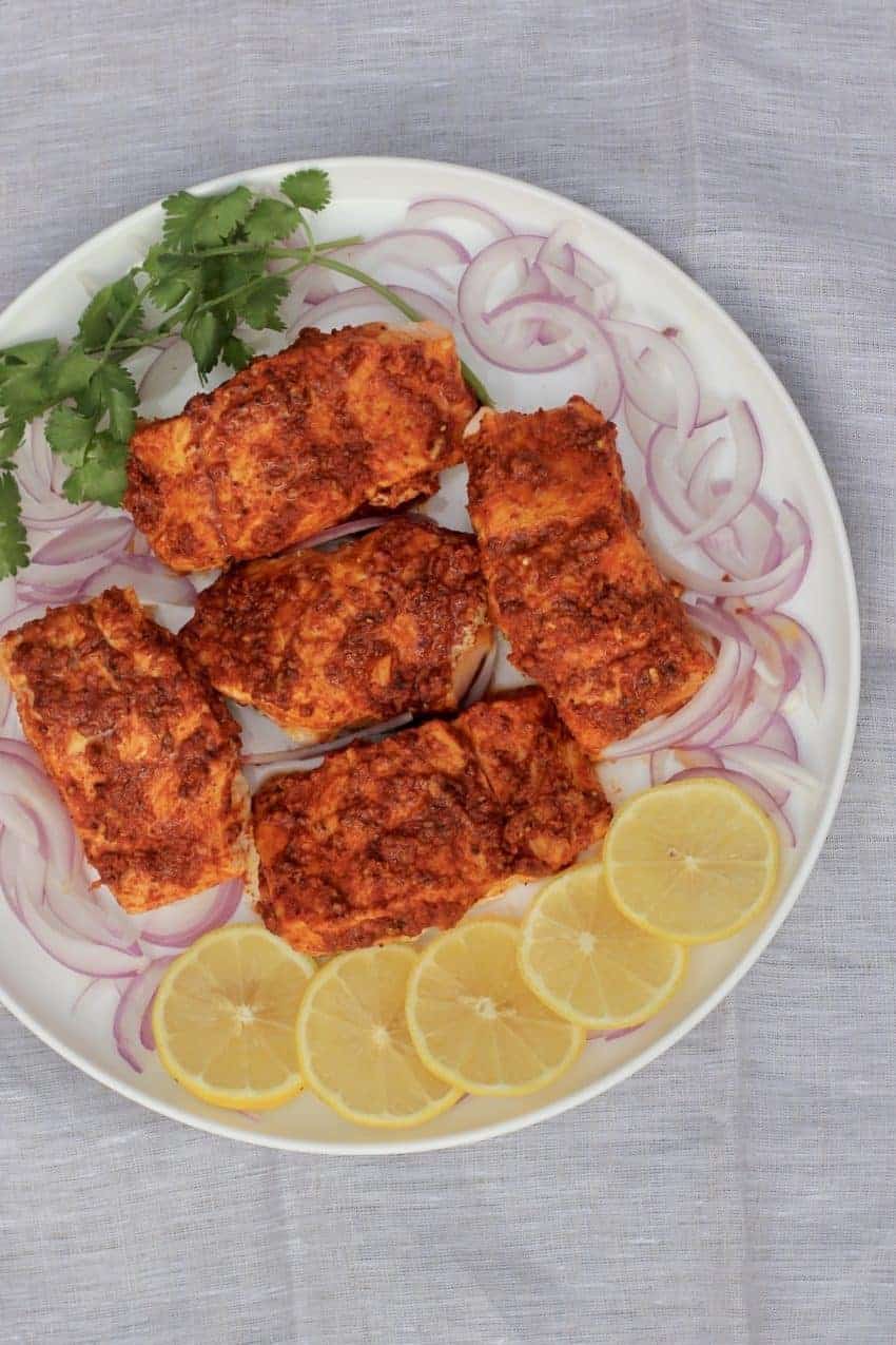 Tandoori Salmon in a white platter with sliced lemons,red onions, and cilantro around the fish.