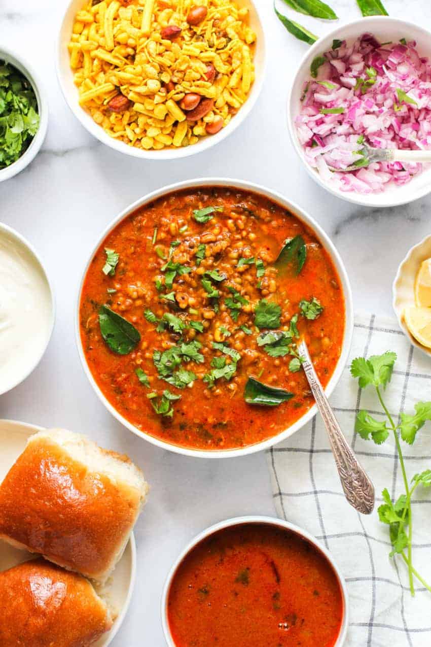 Sprouted bean curry in a white bowl with, dinner rolls and condiments on the side 