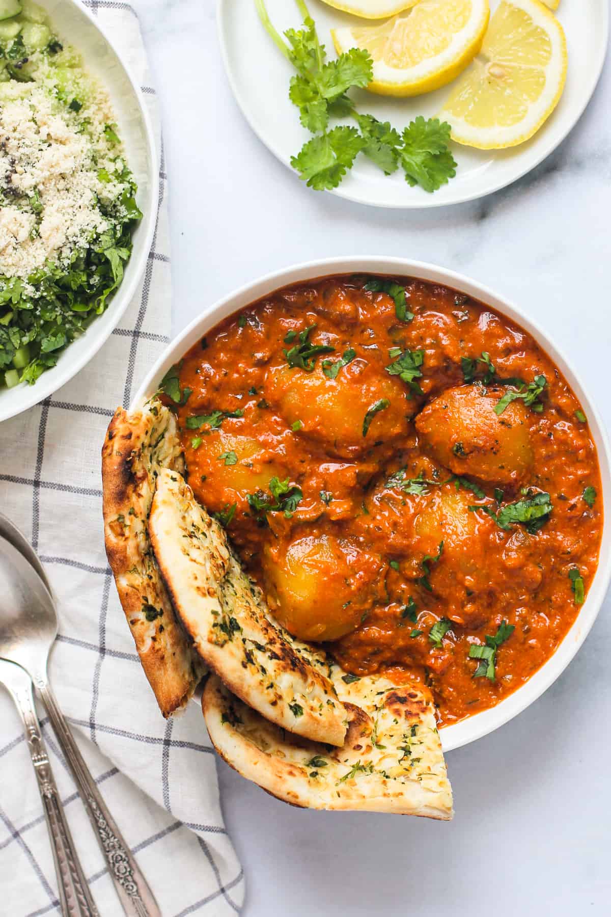 Dum Aloo served with naan in a white bowl with cucumber salad and lemon wedges next to it