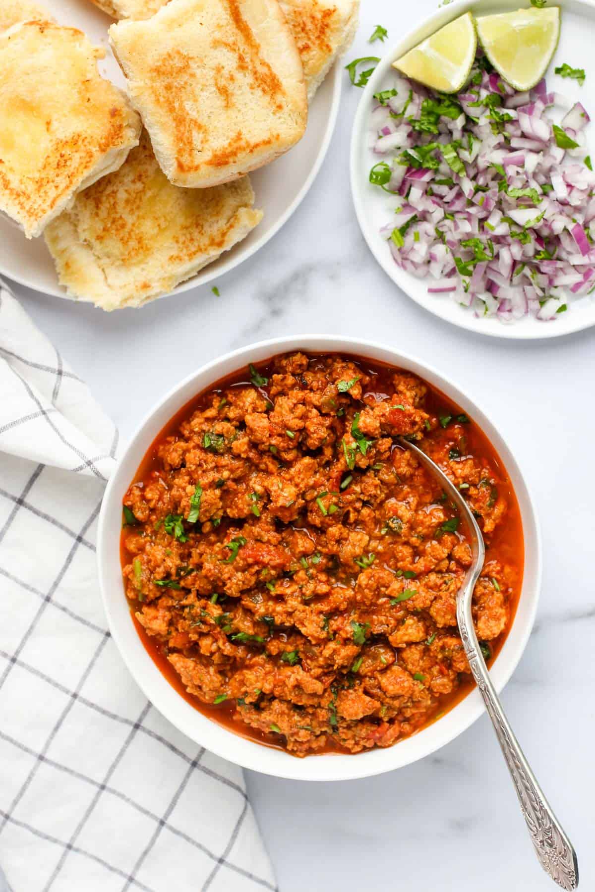 Kheema curry served in a white bowl alongside pav, diced onions and lime 