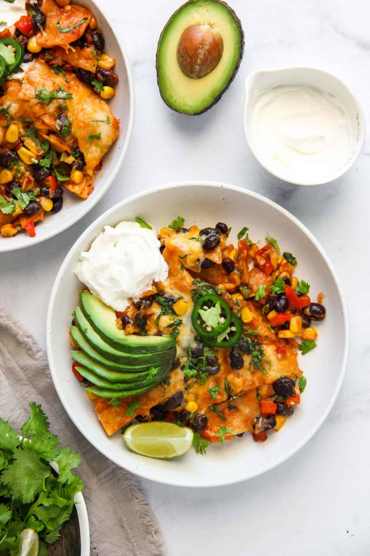 veg enchilada casserole served in a bowl garnished with avocado, sour cream, jalapeños and a lime wedge 