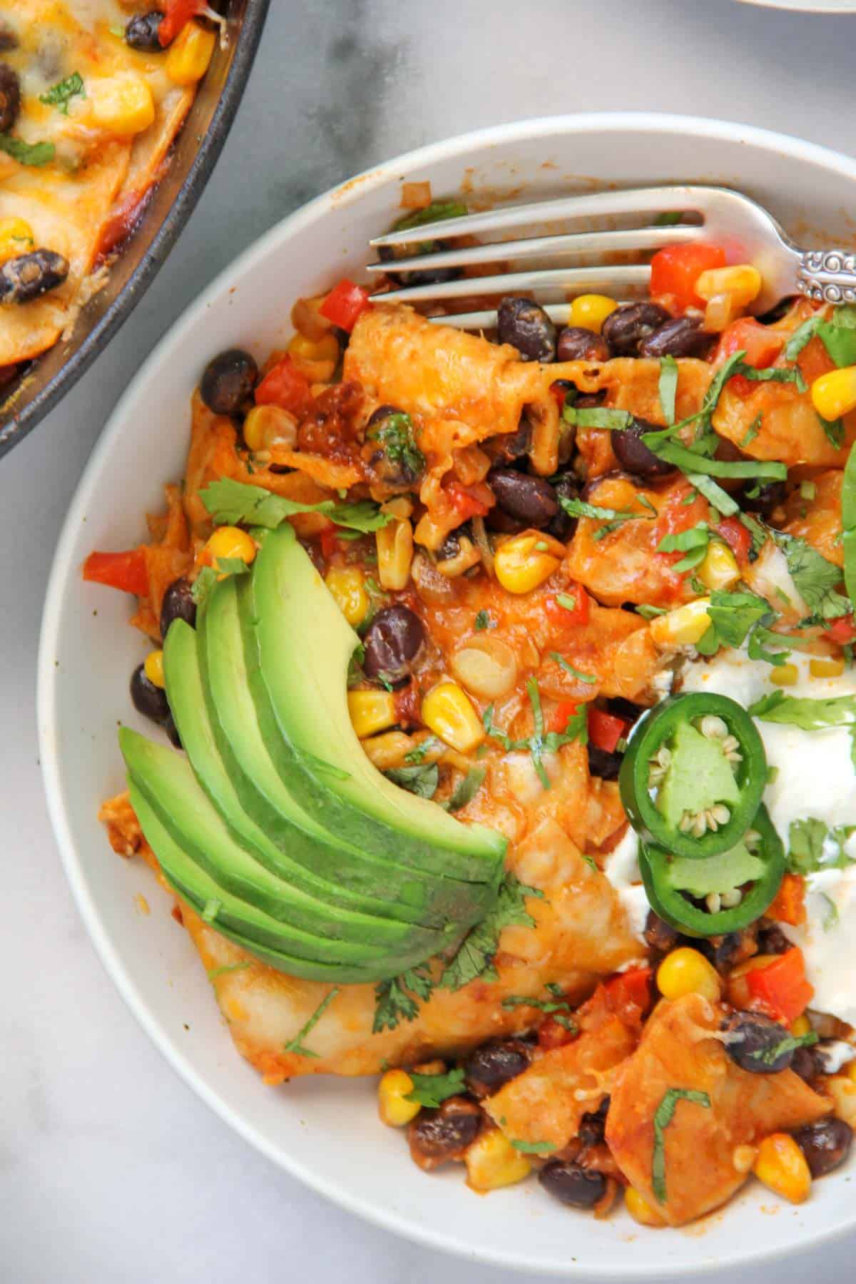 veg enchilada casserole in a white bowl with sliced avocado and jalapeños