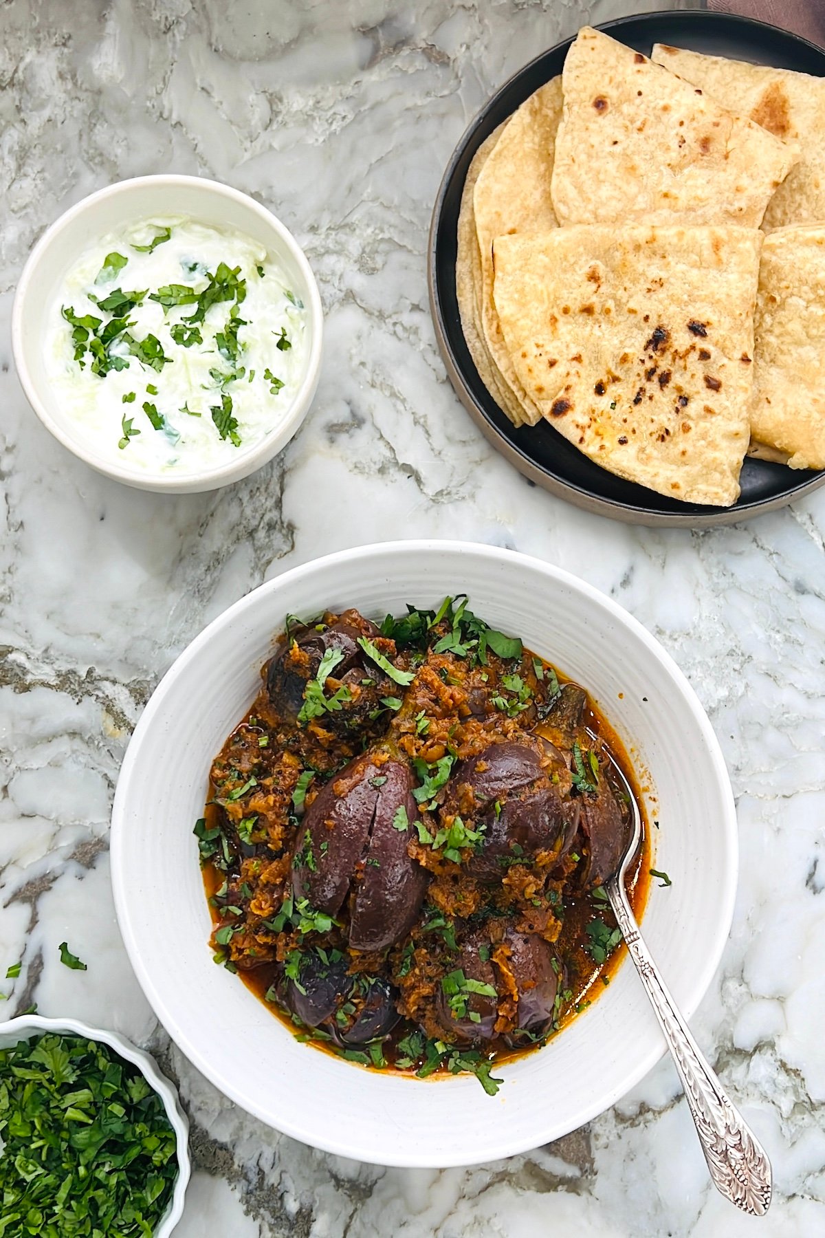 baingan masala in a white bowl with roti and raita