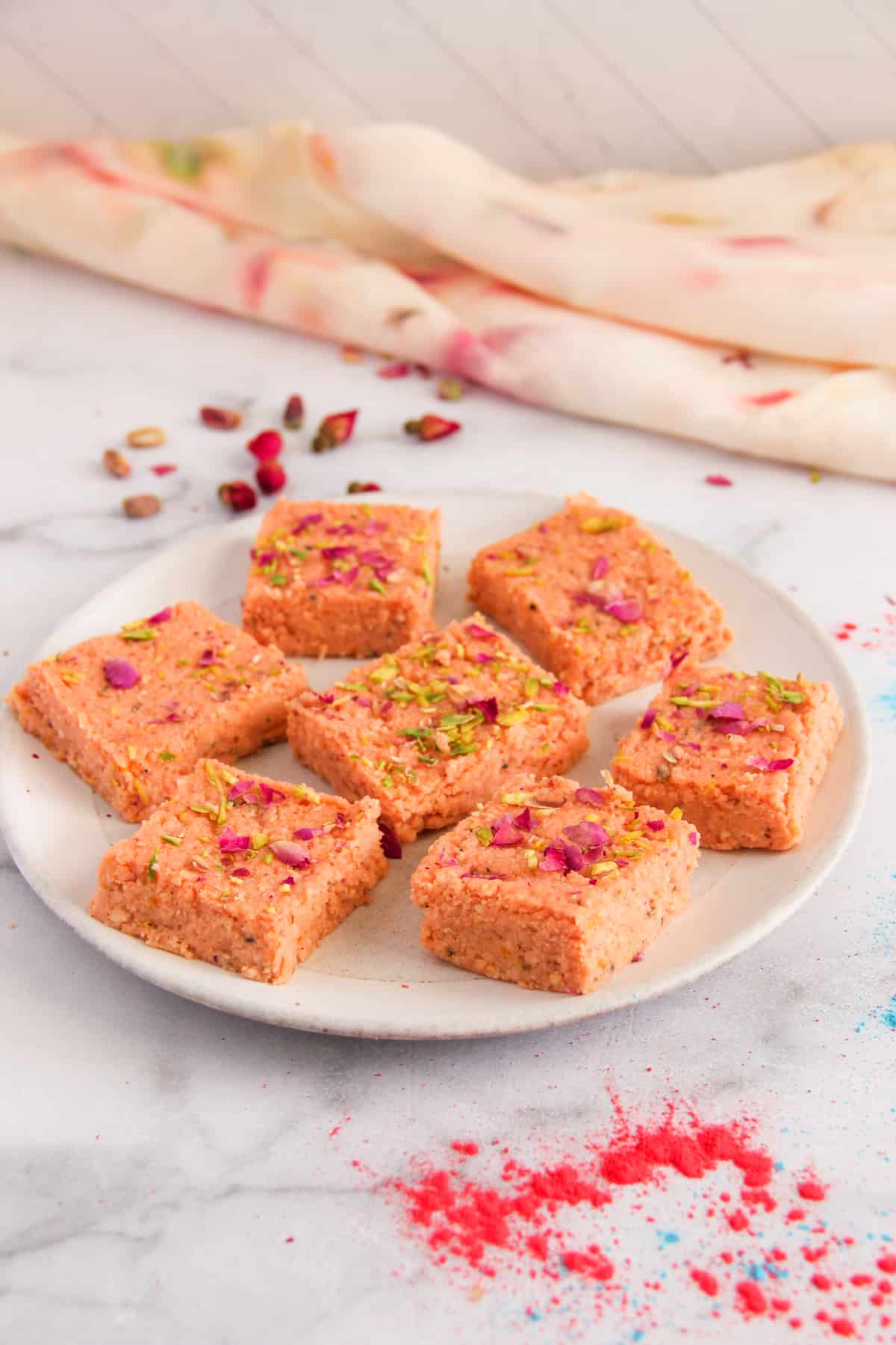 Thandai Kalakand in a white platter garnished with sliced pistachios and rose peatls. 