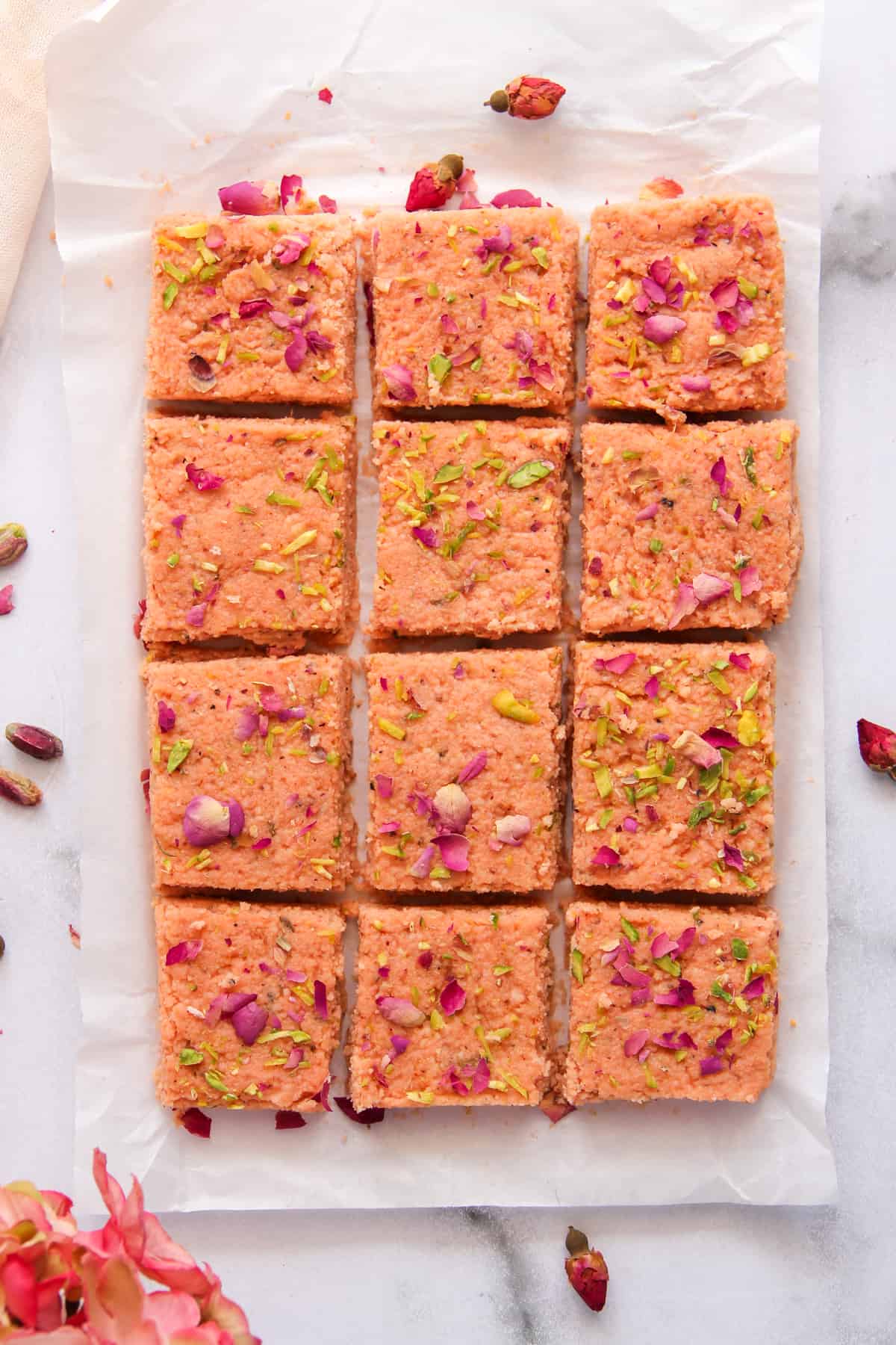 Pieces of thandai kalakand garnoshed with rose petals and pistachios on white parchment paper.
