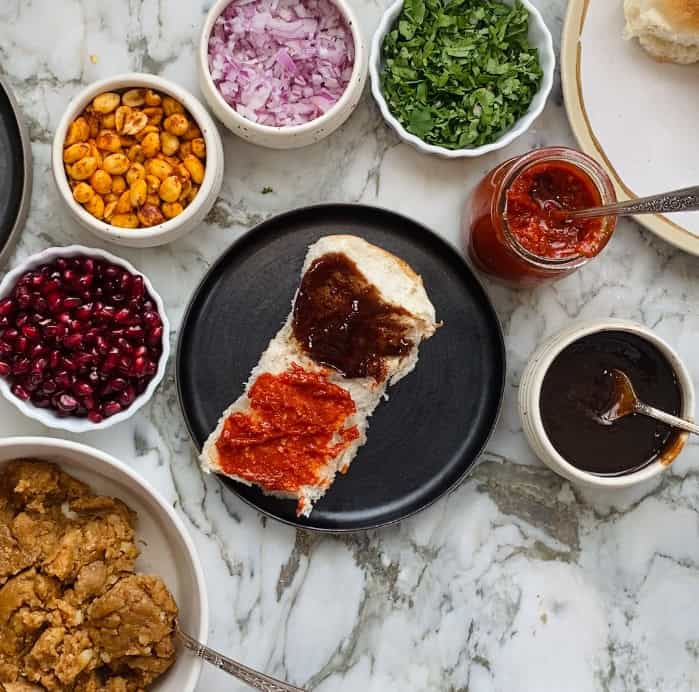 sweet chutney and red chutney on a bun with all the fixings for Dabeli on the side.