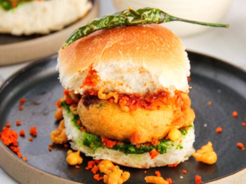 vada pav in a black plate with red chutney, chura and fried chili.