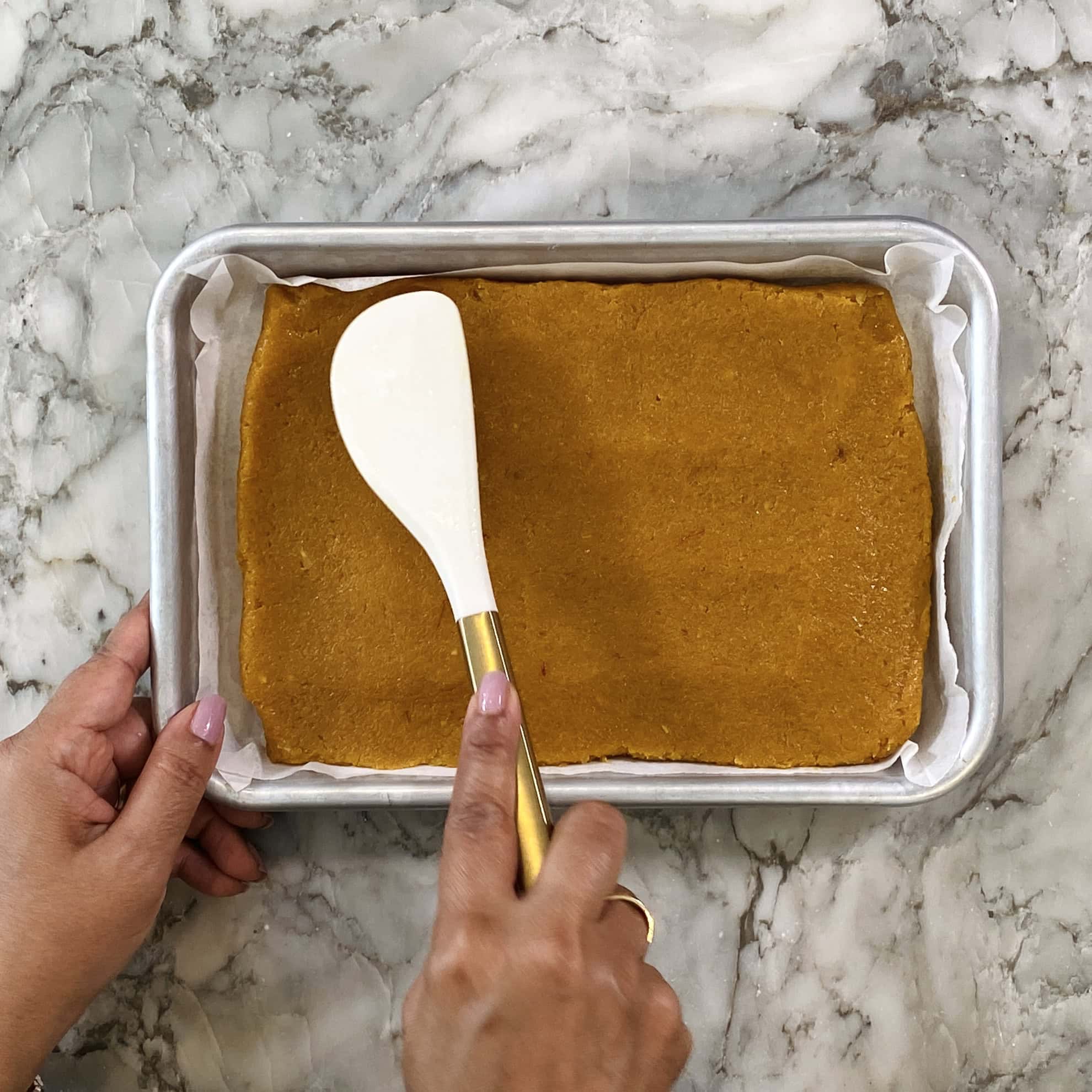 spreading mango coconut burfi dough on a parchment lined tray 